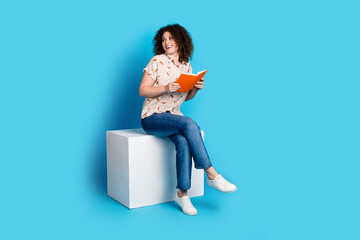 Young woman with curly hair sitting on a white cube reading a vivid orange book against a bright...
