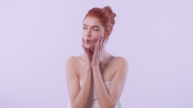 Redhead woman with fair skin playfully touches her face with both hands, showcasing beauty expressions in various angles against soft purple background.