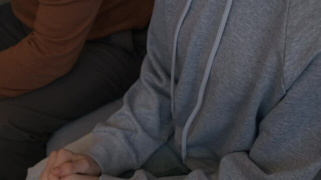 Tilt down shot of teenage Caucasian girl in casualwear and eyeglasses sitting near parent holding hands crossed and feeling vulnerable during family therapy session