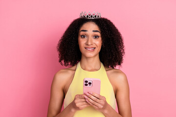 Young woman in yellow dress holding phone, wearing decorative tiara, standing against pink...