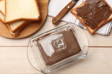 Block of tasty chocolate butter in keeper and slices of bread on light wooden table, flat lay