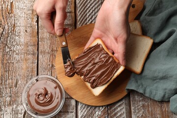Woman spreading tasty chocolate butter onto bread at color wooden table, top view