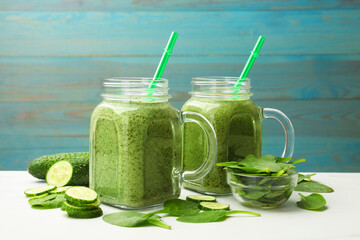 Tasty smoothie in mason jars, spinach leaves and cucumber on white table against light blue wooden background, closeup