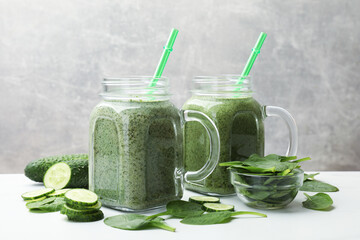 Tasty smoothie in mason jars, spinach leaves and cucumber on white table against grey background,...