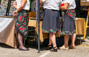 Women in traditional folk skirts and white blouses standing at a market stall during an outdoor...
