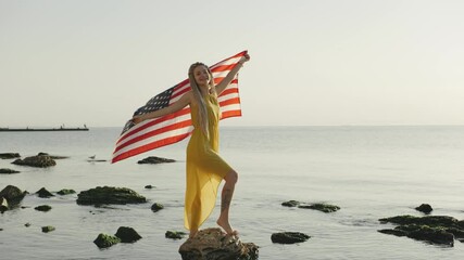 Woman standing on rock in ocean while holding American flag under bright sunlight at a coastal location - Powered by Adobe