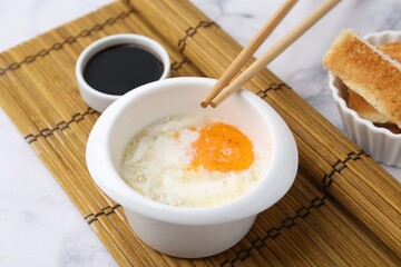 Half-boiled egg in bowl, soy sauce, chopsticks and toasted bread on white marble table, closeup. Traditional asian breakfast