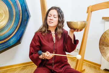 Woman in deep concentration holding a singing bowl, using a mallet for sound healing practices. Her...
