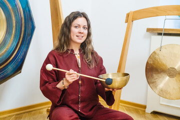 Woman sitting and playing a Tibetan singing bowl, using a mallet to produce calming sounds. She is...