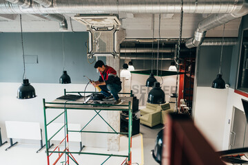 Worker performs electrical maintenance on scaffolding in modern office space during daylight hours