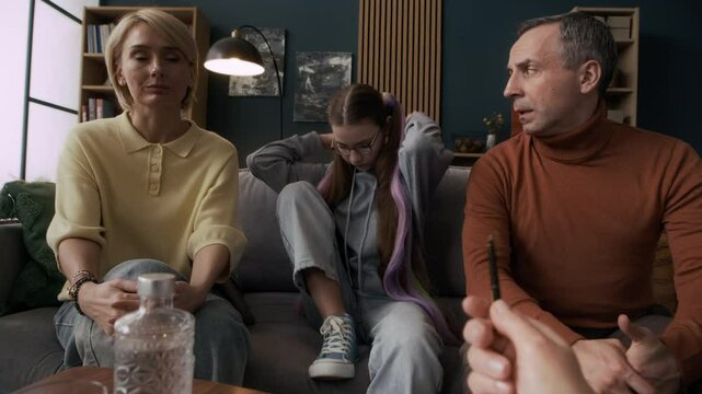 Slowmo of teen Caucasian girl putting on hood and covering her face avoiding conversation with her parents and unrecognizable psychologist during family counseling session in modern office