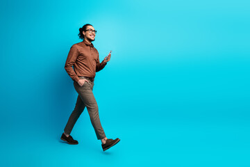 Confident young businessman walking while using smartphone against vibrant blue background