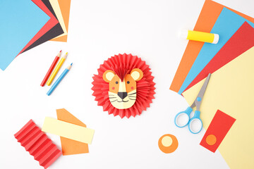Handmade paper lion head and craft supplies on a white background. Flat lay composition with scissors, glue stick, colored paper, and cut-out shapes. DIY kids craft and creative hobby concept,
