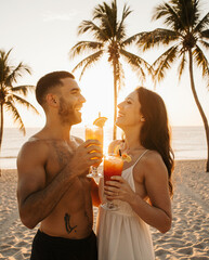 A mixed-race couple (Latino man and Caucasian woman) laughing on a beach at sunset with tropical cocktails. Palm trees and ocean in the background.