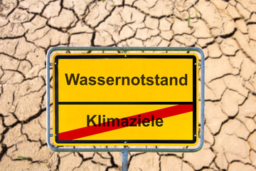 Vor ausgetrocknetem Boden mit vielen Rissen steht symbolisch ein Schild mit dem Aufdruck...
