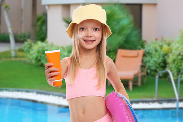 Cute little girl with tube of sunscreen and inflatable ring by outdoor swimming pool. Sun...