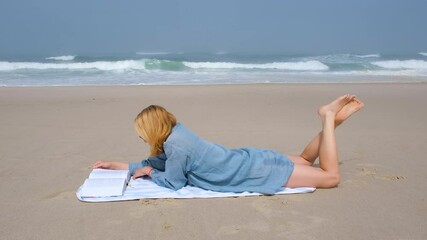 A young woman lies on her stomach on a beach towel, reading a book by the Atlantic Ocean. She enjoys a peaceful, sunny day with waves crashing gently in the background