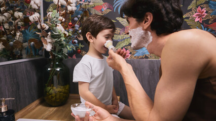 In a vibrant bathroom, a dad playfully dabbed shaving cream onto his young son's nose. Concept of father-child emotional connection, involved fatherhood