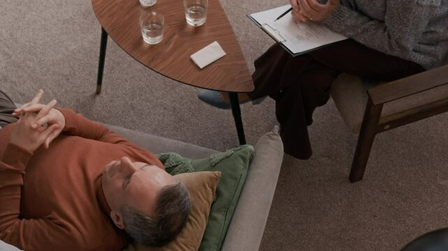 High angle shot of middle-aged Caucasian male patient lying down on couch and starting individual therapy session with unrecognizable psychologist holding notes