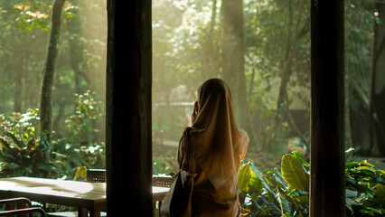 Woman in hijab gazes at sunlit tropical forest from indoors, a serene and contemplative moment with...