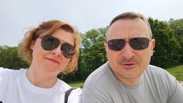 Happy middle-aged couple wearing sunglasses taking a selfie outdoors on a sunny day, smiling together in a green park environment