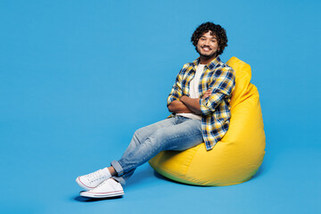 Full body young smiling happy Indian man wear yellow checkered shirt casual clothes sit in bag...
