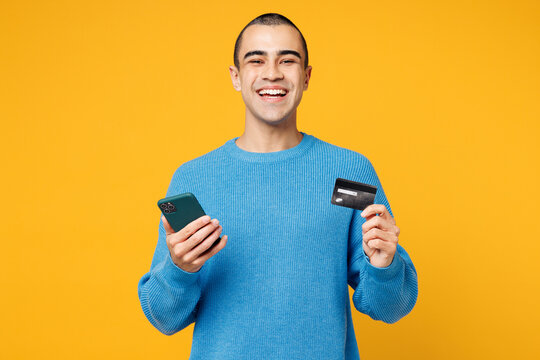 Young middle eastern man wearing blue sweatshirt casual clothes using mobile cell phone hold credit bank card do online shopping order delivery booking tour isolated on plain yellow orange background.