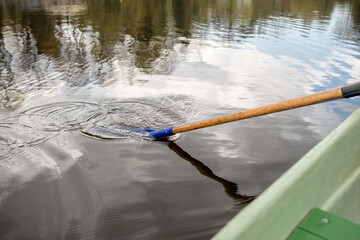 A person paddles a wooden oar through serene water, creating ripples while surrounded by trees and a cloudy sky during a peaceful day.