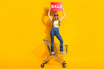 Smiling woman holding sale sign while standing in shopping cart on vibrant yellow background...
