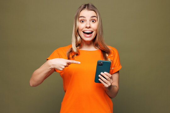 Young shocked surprised woman wears orange t-shirt casual clothes hold in hand use point index finger on mobile cell phone isolated on plain pastel green background studio portrait. Lifestyle concept.
