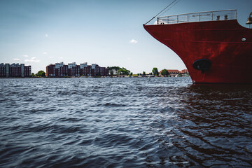In light waves, a red ship's hull protrudes into the harbor basin against an urban backdrop,...