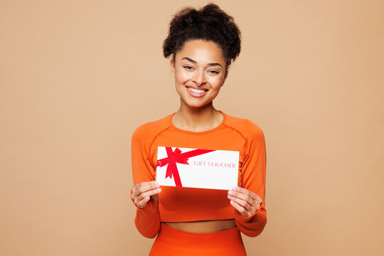 Young strong fitness trainer instructor sporty woman wear orange clothes spend time in home gym hold store gift coupon voucher card isolated on plain beige background. Workout sport fit abs concept.