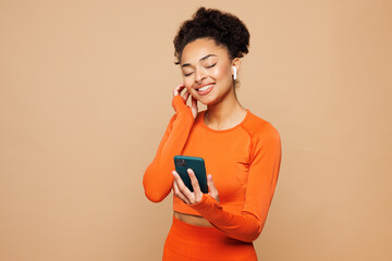 Young fitness trainer instructor sporty woman wear earphones orange clothes spend time in gym hold use mobile cell phone listen music isolated on plain beige background. Workout sport fit abs concept.