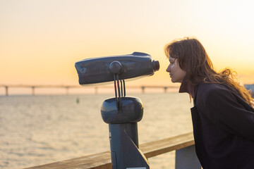 Girl looks through public stationary binoculars on the river bank, coin operated metal binoculars.
