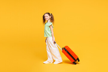 Side view traveler kid child girl in green casual clothes hold suitcase bag isolated on plain yellow background. Tourist travel abroad in free spare time rest getaway Air flight trip journey concept
