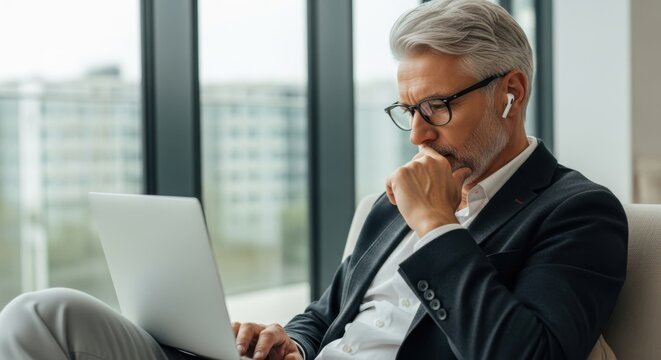 Mature businessman wearing glasses and suit using laptop and earbuds in modern office - Powered by Adobe