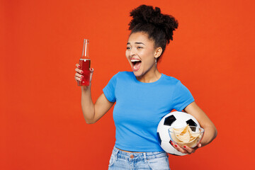 Young overjoyed happy woman fan wears basic blue t-shirt cheer up support football sport team hold in hand soccer ball bottle drink beer watch tv live stream isolated on plain red orange background.
