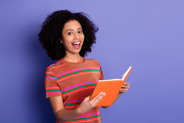 Smiling young woman holding an orange book, wearing a colorful t-shirt against a vibrant purple...