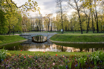 A serene park features a picturesque bridge crossing a tranquil river, bordered by colorful flowers...