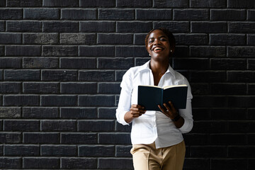 Portrait of elegant black African woman leaning on the brick wall and holding book.
