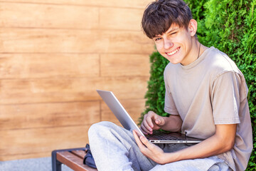 Happy teenage boy with laptop sitting on the street, portrait of student with laptop