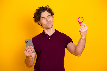 Young man with curly hair holding a smartphone and an object, standing against a bright yellow...