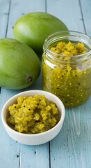 Three green mangoes rest beside a jar and bowl of tangy mango chutney on a blue wooden surface—homestyle ingredients infused with texture and heritage.