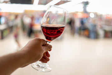 Close-up of a hand holding a glass of red wine against a blurred indoor background, evoking elegance, leisure, and refined lifestyle in a casual public setting.