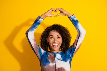 Vibrant young woman creating a heart shape gesture while smiling brightly against a cheerful yellow...