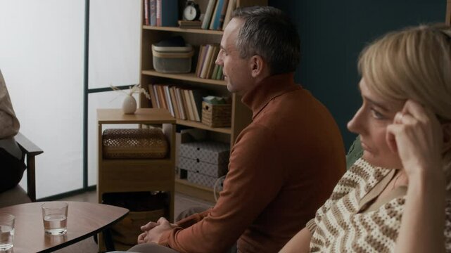 Slowmo shot of middle-aged Caucasian man talking to mature female counselor during couples therapy while his wife turning away avoiding conversation and looking frustrated