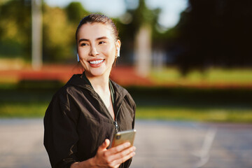 Smiling sporty woman listening music using wireless earbuds and holding smartphone in a park