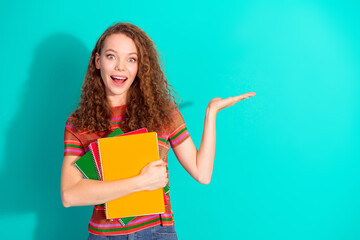 Smiling young girl in a casual striped t-shirt holding colorful notebooks on a vibrant teal...