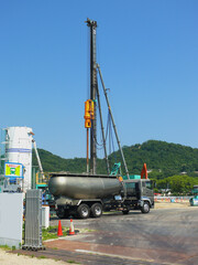 A landscape view of the construction site from the entrance. A cement storage tank stands beside a cement truck, with a pile driver visible behind them.