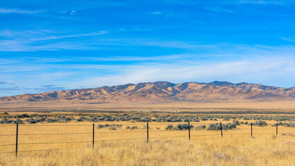 Barbed wire fence crossing dry grassland in Nevada desert landscape with mountains and blue sky